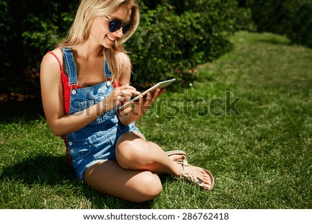 Portrait of happy young blonde woman college student use tablet pc sitting on grass in park.