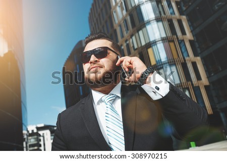 Portrait of successful business man talking on smart phone outside office. Attractive adult man in suit talking on the phone and standing near his financial office  background, flare light