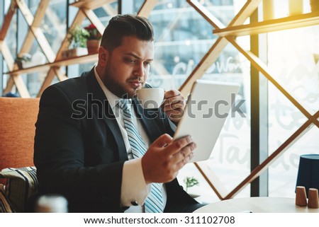 Portrait of successful business man drinking coffee inside coffee shop. Attractive adult man in suit looking on digital tablet pc, flare light
