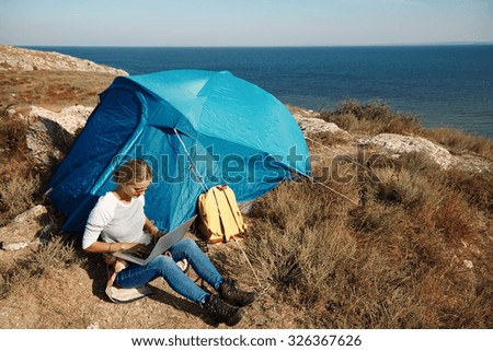 Happy woman sitting near tent with laptop, view of mountains ,sky and sea.