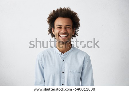 Photo of Handsome unshaven young dark-skinned male laughing out loud at funny meme he found on internet, smiling broadly, showing his white straight teeth. Positive human facial expressions and emotions