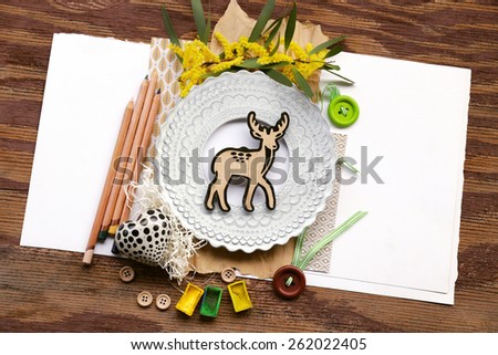 Spring nature composition with baby deer placed over shredded paper next to pencils and artistic white photo frame with buttons over white placeholder paper on the left and right and vintage wood