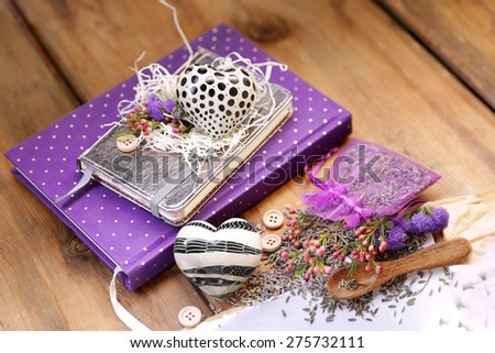 Artistic composition with lavender seeds and flowers next to a purple diary notepad under a silver book and heart next to an organza bag, wooden spoon and a sheet of paper on brown wood