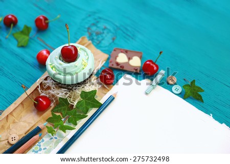 Bright composition with a bunch of ripe red cherries and a chocolate muffin placed next to a piece of milk chocolate over brown vintage paper next to a pencil and green leaves on blue textured wood