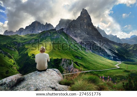 The young man is resting on the rock after hiking trip, he is looking to green mountains meadows under towering hill in italian Dolomite.