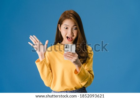 Surprised young Asia lady using mobile phone with positive expression, smiles broadly, dressed in casual clothing and standing isolated on blue background. Happy adorable glad woman rejoices success.
