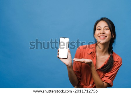 Young Asia lady show empty smartphone screen with positive expression, smiles broadly, dressed in casual clothing feeling happiness on blue background. Mobile phone with white screen in female hand. 商業照片 © 