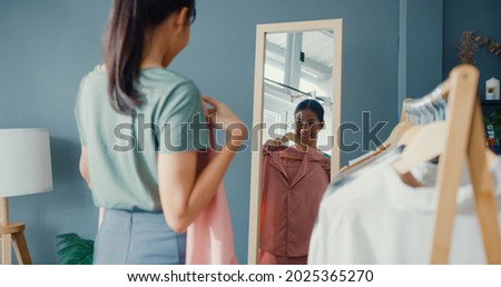 Beautiful attractive Asia lady choosing clothes on clothes rack dressing looking herself in mirror in living room at house. Girl think what to wear casual shirt. Lifestyle women relax at home concept.