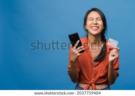 Young Asia lady using phone and credit bank card with positive expression, smiles broadly, dressed in casual clothing and stand isolated on blue background. Happy adorable glad woman rejoices success.