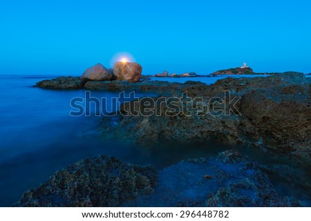 Gold hour over the wild rocky beach coastline and the sea. Lighthouse, moon and rocks at night wide angle view. Panoramic landscape in Sardegna, Nora place near Pula city.