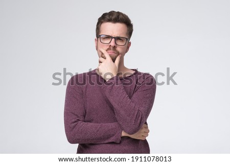 Generating new ideas for you. Thoughtful young man holding hand on chin and looking at camera while standing isolated on white wall
