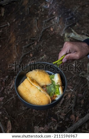 man's hand holding a pot with bread sandwich with lettuce and tomato inside in a camping context