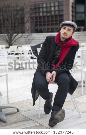 young sad man wearing check cap and red scarf  and red scarf sitting at the street cafe waiting for order