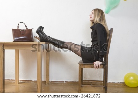 young blond woman sitting on chair at table put her legs on table. Bag standing on table. Room decorated balloons.