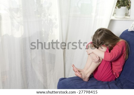 Little cute dreaming girl sitting on sofa at window hugging her knees. Child`s grief