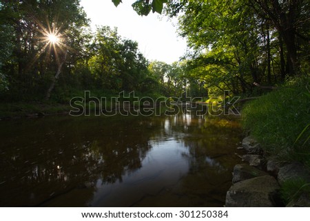 Sun setting through trees lining a shallow rocky creek. Taken at sundown and at the peak of summer. Long exposure with super wide perspective.