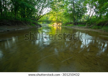 Super wide shot of a shallow stream flowing towards a bridge in the distance. Image taken at sundown during the height of summer.