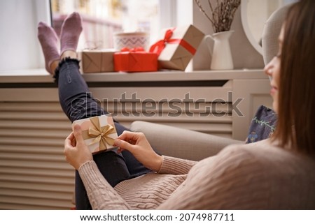 PRETTY young woman while sitting in a comfortable armchair holding a gift box surrounded by gifts at home. Ghe guesses what's inside Imagine de stoc © 