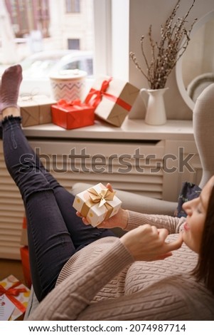 PRETTY young woman while sitting in a comfortable armchair holding a gift box surrounded by gifts at home. Ghe guesses what's inside Imagine de stoc © 