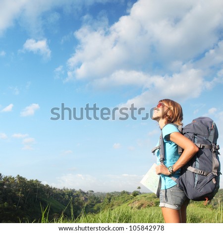 Young smiling woman standing on a green meadow with map and backpack and looking to a blue sky