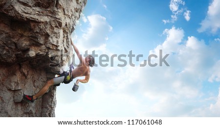 Young man climbing vertical wall with belay with blue cloudy sky on the background