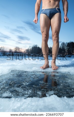 Young man standing by ice hole and ready to swim in the winter water