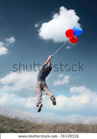 Young man flying in blue sky over meadow with grass holding colored balloons.