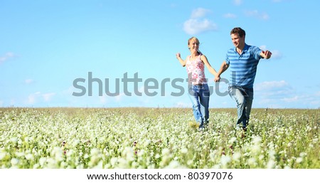 Young happy man and woman running on meadow with fluffy dandelions on blue sky background