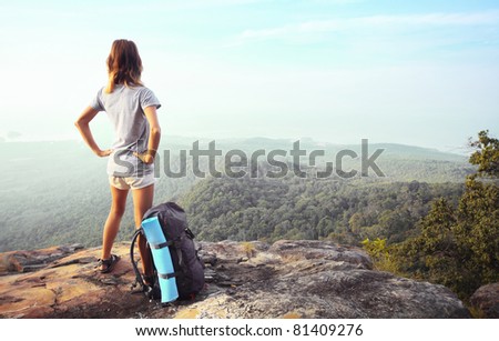 Young woman standing with backpack on cliff\'s edge and looking into a wide valley