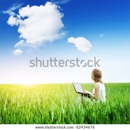 Young woman with laptop sitting on green meadow and looking to a sky with rare clouds