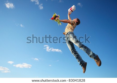 Young man jumping with flowers and gift box on blue sky background