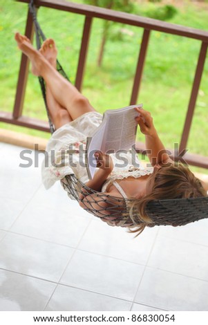 Young woman lying in a hammock in a garden and reading a book