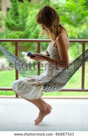 Young woman sitting in a hammock in a garden and reading a book