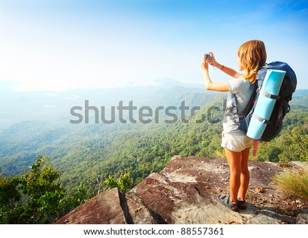 Young woman standing with backpack on cliff's edge and taking a photo of a wide valley