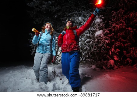 Women with backpacks exploring deep wild forest at night with a torch and red flare