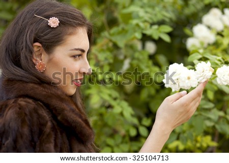 A close up portrait of a rich beautiful woman in natural brown fur and with rose accessories in her long wavy hair with white roses on background. Autumn.