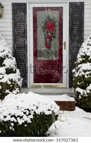 Cape-cod style home with front red door and wreath for the holidays in the midst of a snowstorm.