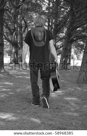 Black and white image of Guitarist walking while carrying his guitar to the side in wooded area.