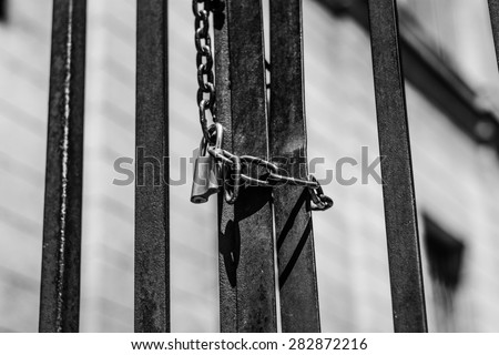 Close up photo of a padlock with big thick chain lock on a black metal gate. 
Padlock was photographed outside in Szeged, Hungary.The gate was locked. Selective focus.