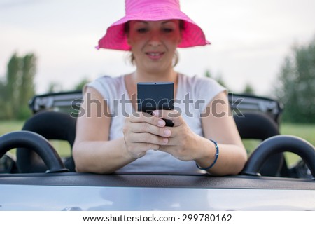 Young attractive girl using mobile phone in her car with the smile on her face. She is wearing pink hat and white shirt. Mobile phone is black, while coupe cabriolet car is bright blue.
