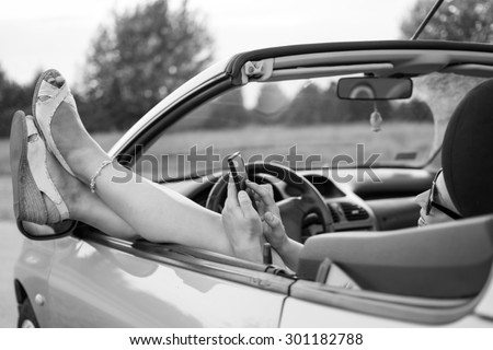 Young attractive girl using mobile phone in her car.She is wearing black glasses and white sandals.Mobile phone is black,while coupe cabriolet is bright blue. Focus on glasses.Black and white photo.
