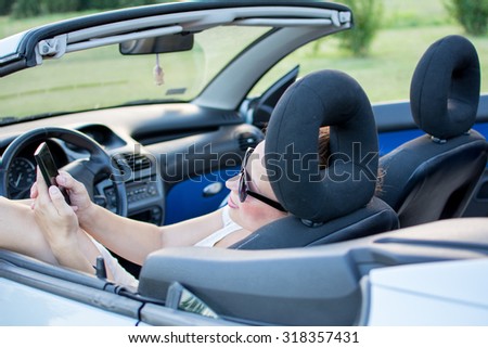 Young attractive girl sitting in convertible car and holding mobile phone.She is wearing black glasses and white shirt. Mobile phone is black, while convertible car is bright blue.
