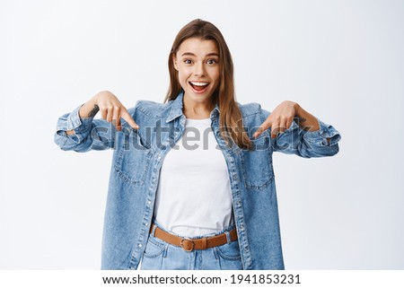 Excited blond woman checking out awesome news, pointing fingers down and staring amazed, showing advertisement on bottom copy space, standing against white background
