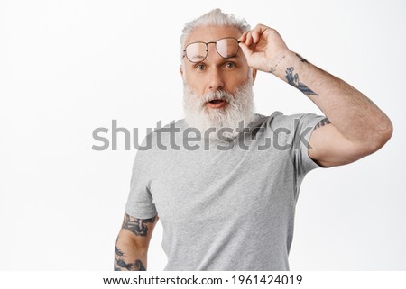 Surprised mature guy with long beard, take-off glasses and look in awe, staring at something amazing, standing in grey t-shirt against white background Foto stock © 