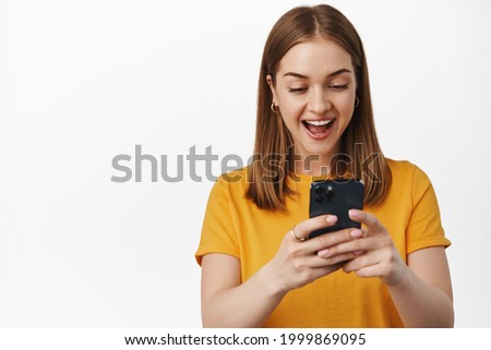 Portrait of girl looks surprised and excited at smartphone screen, receive pleasant notification, watching video on mobile phone, cellular internet, white background