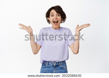 Image of excited and surprised brunette woman gasp fascinated, spread hands sideways and looking speechless impressed, rejoicing, being happy for someone, white background Foto stock © 