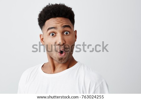 Close up of funny handsome young dark-skinned man with afro haircut in stylish white t-shirt looking in camera with raised eyebrows and surprised face expression. Stockfoto © 