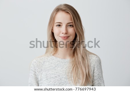 Photo of Pretty smiling joyfully female with fair hair, dressed casually, looking with satisfaction at camera, being happy. Studio shot of good-looking beautiful woman isolated against blank studio wall.
