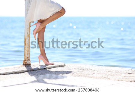 woman in white dress leaning standing by the sea side looking at the sea with stilettos