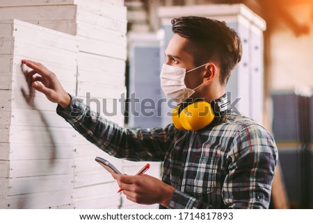Portrait male foreman inspector in medical face mask and protective headphones checking woodwork stock at factory storage. Man supervisor counting wood inventory. Warehouse worker. COVID-19 quarantine Photo stock © 